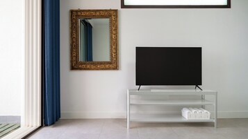 Una habitación con una televisión sobre un soporte blanco, un espejo con marco dorado en la pared y una ventana con cortinas azules.