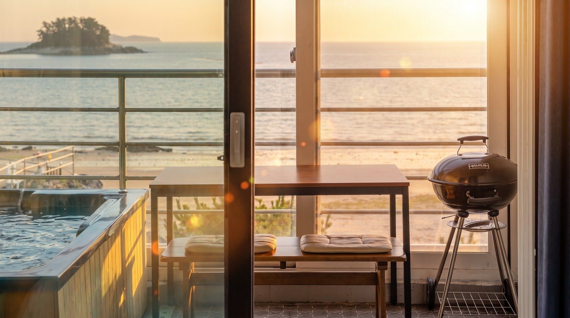 A balcony with a view of the ocean, a hot tub, a table with cushions, and a barbecue grill.