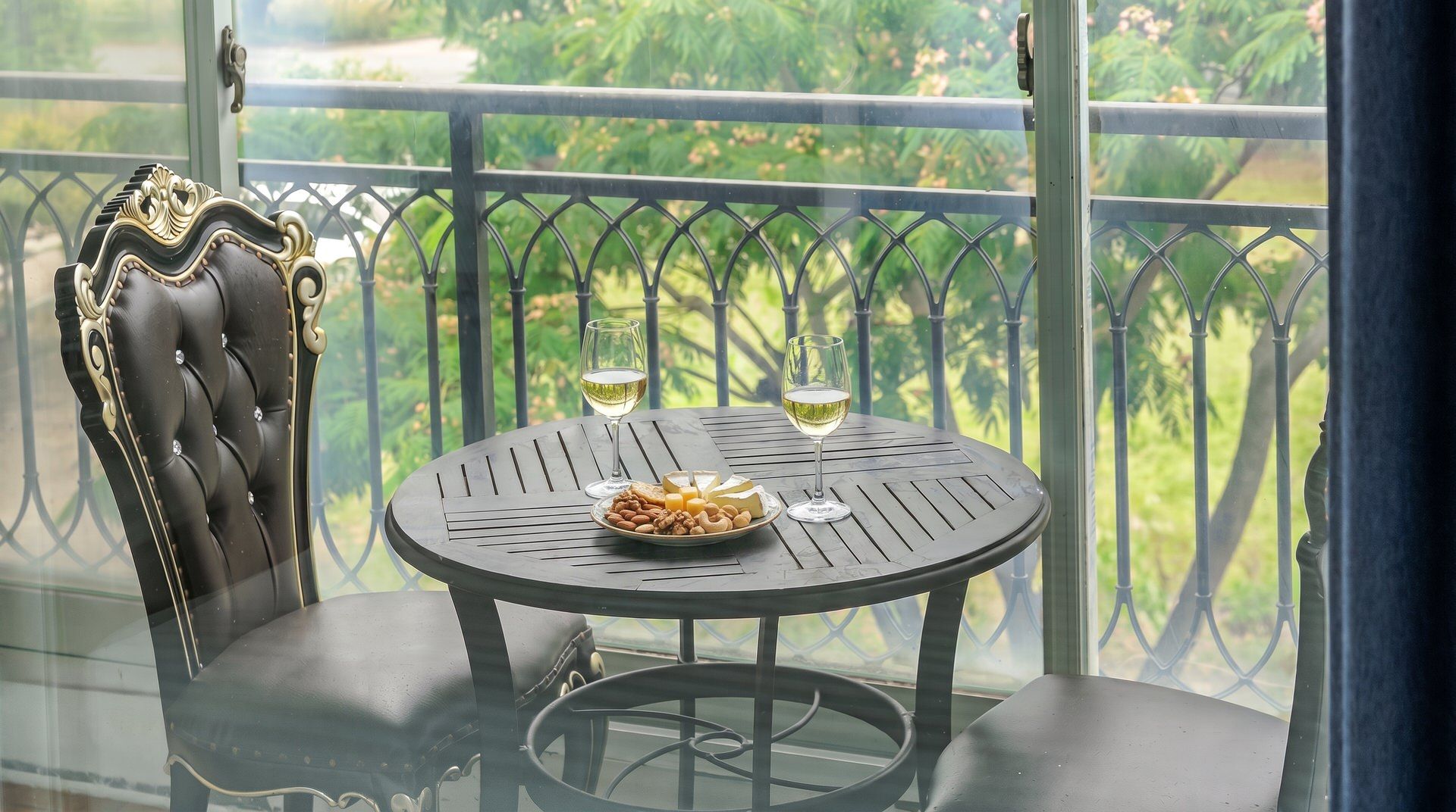 A round table with two glasses of white wine, a plate of cheese and nuts, and a black upholstered chair with ornate detailing.