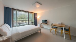A hotel room with a bed, a desk with a chair, a TV, and a balcony with a view of trees. - The Seaside Taean (Taean)