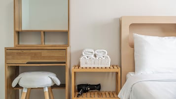 Habitación de hotel con una cama, una mesita de noche de madera, un taburete pequeño, un espejo y un estante con toallas y un objeto negro.