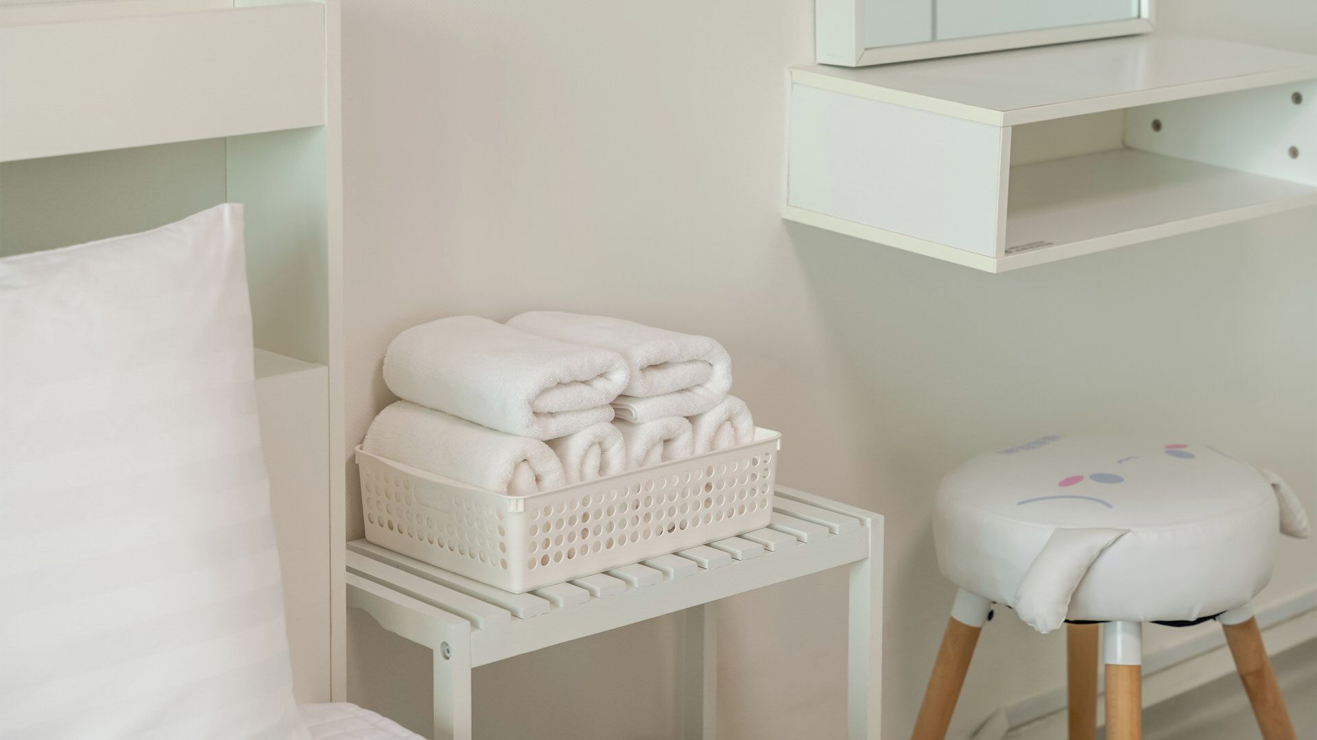A white room with a bed, a small white stool, a basket of folded towels, and a shelf with a mirror.