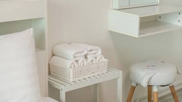A white room with a bed, a small white stool, a basket of folded towels, and a shelf with a mirror.