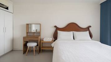 A hotel room with a bed, a wooden desk with a mirror, a small stool, and a shelf with toiletries.