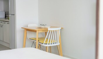 A small, neatly arranged room with a single bed, a wooden table, and a white chair.