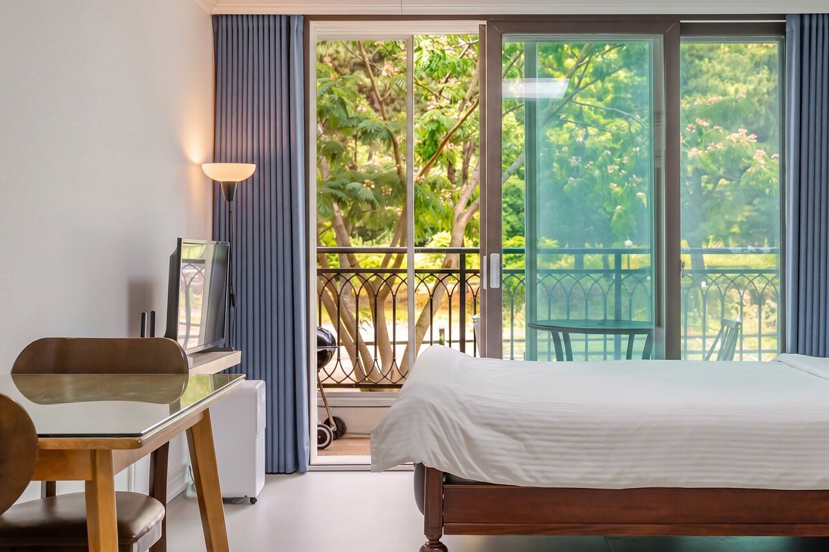 A bedroom with a bed, a desk, and a chair. There is a balcony with a view of trees and a railing.
