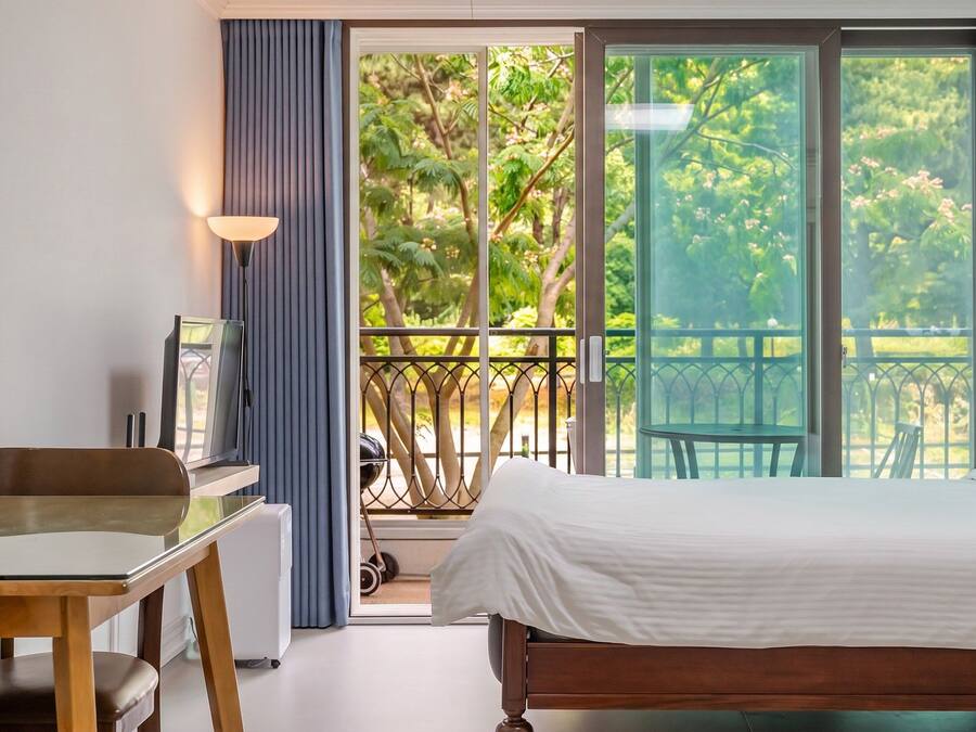 A bedroom with a bed, a desk, and a chair. There is a balcony with a view of trees and a railing.