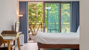 A bedroom with a bed, a desk, and a chair. There is a balcony with a view of trees and a railing.
