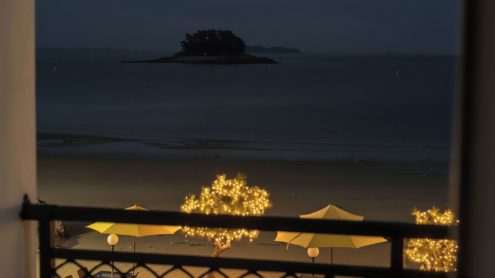 Vista da praia com uma ilha distante, guarda-sóis e luzes.