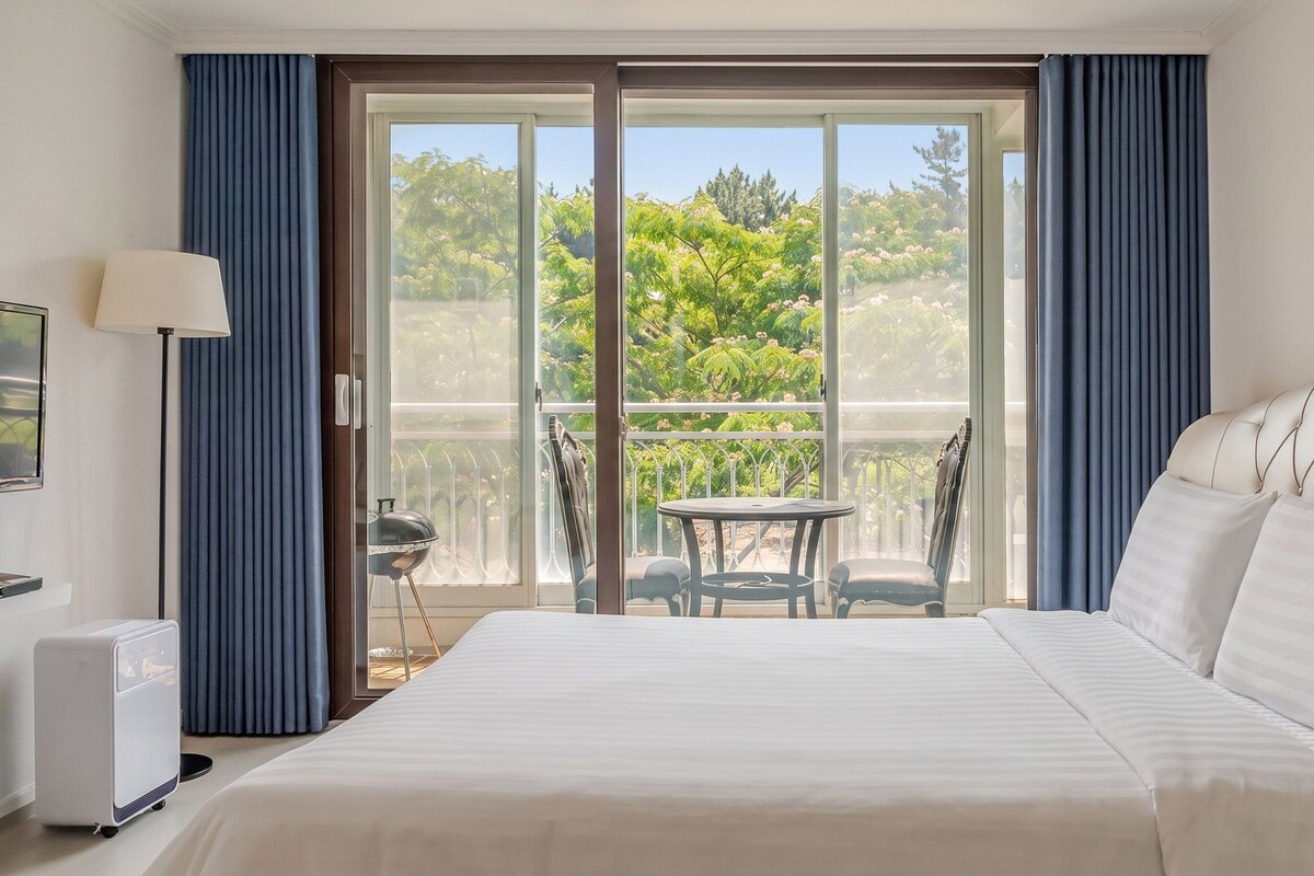 A hotel room with a large bed, a table, chairs, and a view of greenery through a sliding glass door.