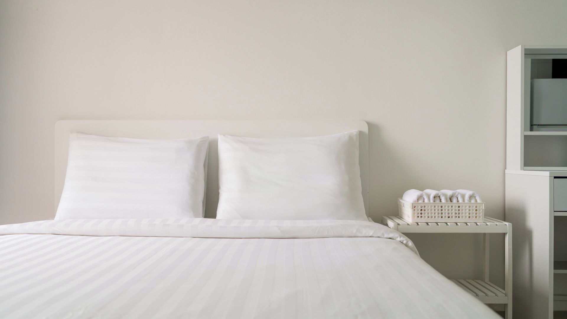A bed with white bedding, a white nightstand with a basket of towels, and a white wall.