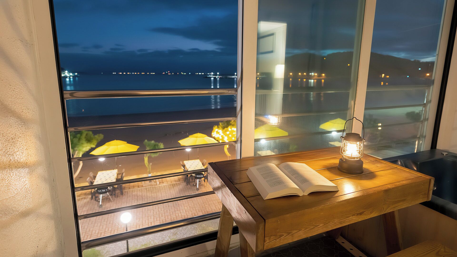 A wooden table with an open book and a lantern, positioned by a window overlooking a cityscape at night.