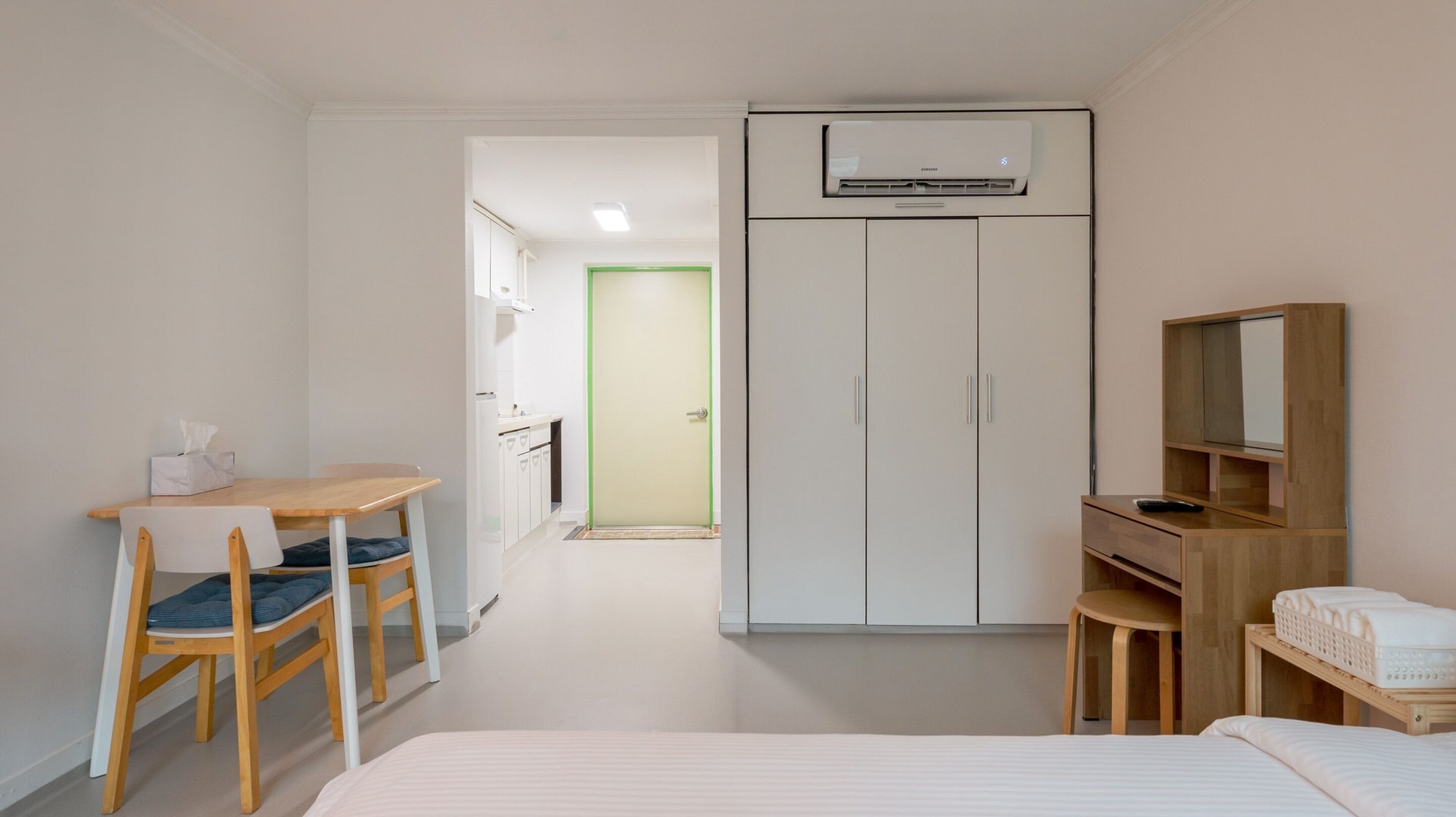 Une chambre d’hôtel de taille réduite comprenant un lit, un bureau avec une chaise, une armoire et un petit coin salon avec une table et des chaises.
