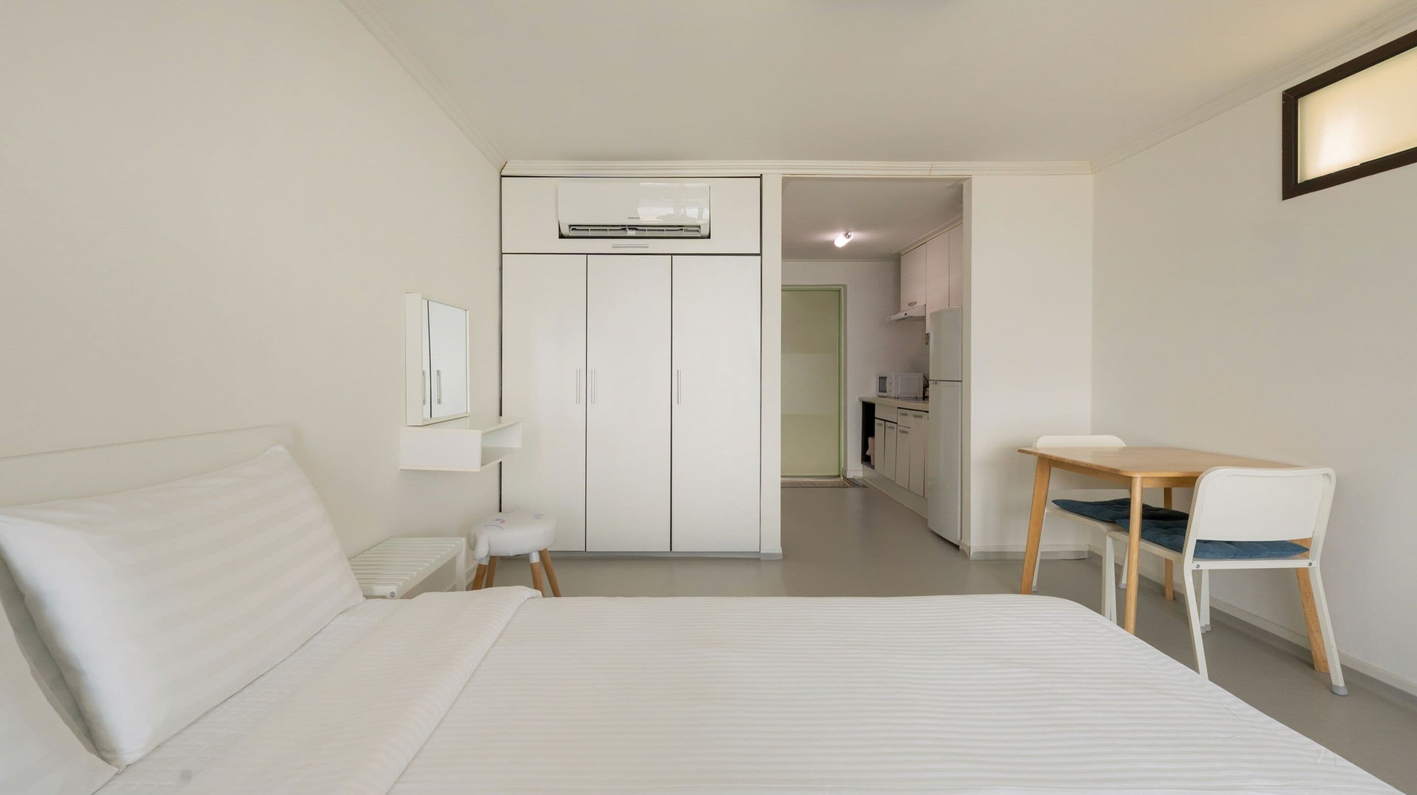 Habitación de hotel con cama, escritorio, silla, una mesita, espejo y una kitchenette al fondo.