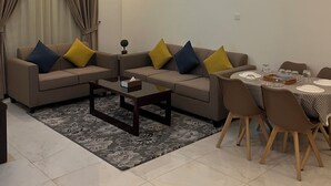 A modern living room with a sofa set, coffee table, dining table, and a painting on the wall. - Danat Al Sharq Serviced Apartments (Al Khobar)