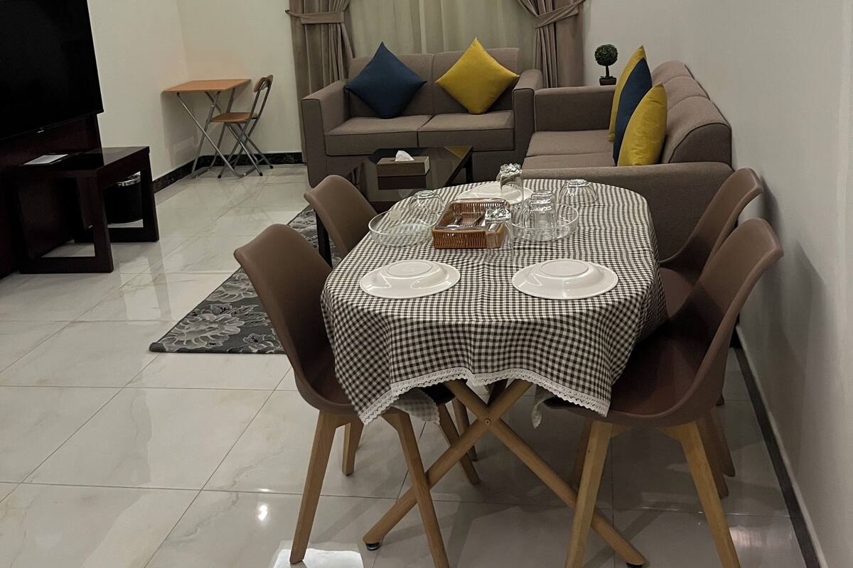 A modern living room with a dining table, chairs, a sofa with cushions, a TV, and a small table with a chair.