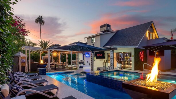 Area piscina con poltrone prendisole, un elemento decorativo a fuoco e un edificio con il cartello 'NETFLIX'.