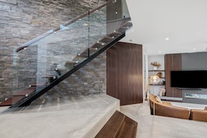 A modern living area with a stone wall, a glass staircase, a flat-screen TV, and a brown leather sofa. - Stonewood by Coast Hosting (North Avoca)
