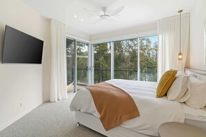 A modern bedroom with a large bed, a ceiling fan, a TV mounted on the wall, and a sliding glass door leading to a balcony with a view of trees. - Stonewood by Coast Hosting (North Avoca)