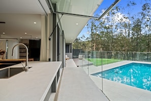 Una cocina moderna con una isla grande, una piscina exterior cubierta con vidrio y vistas a los árboles circundantes.