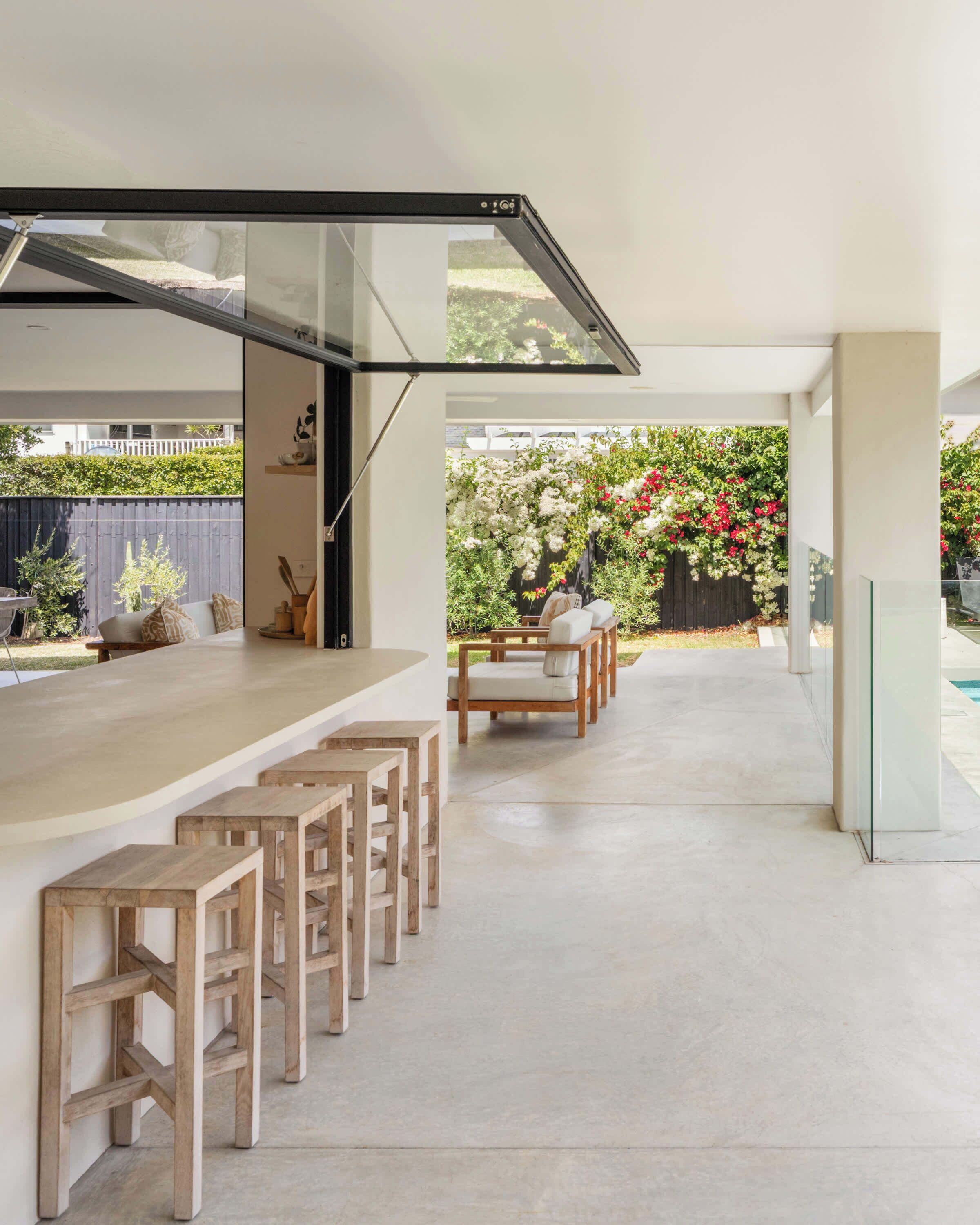 Uma área de estar moderna ao ar livre com bancos de madeira e uma mesa com tampo de vidro, rodeada por um jardim com plantas floridas.