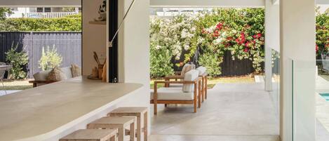 Un espace de détente extérieur moderne, doté de tabourets en bois et d’une table à plateau en verre, entouré d’un jardin planté de fleurs.