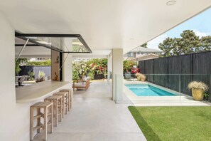 Una cocina moderna al aire libre con barra, piscina y césped bien cuidado.