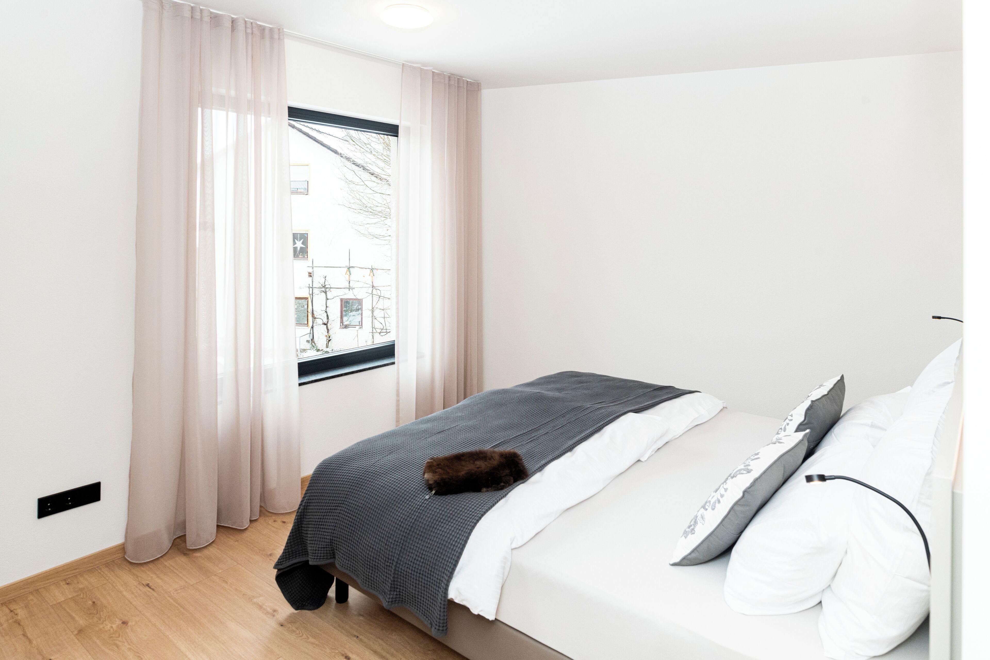 Un dormitorio minimalista con cama, ventana con cortinas translúcidas y vista a un edificio.