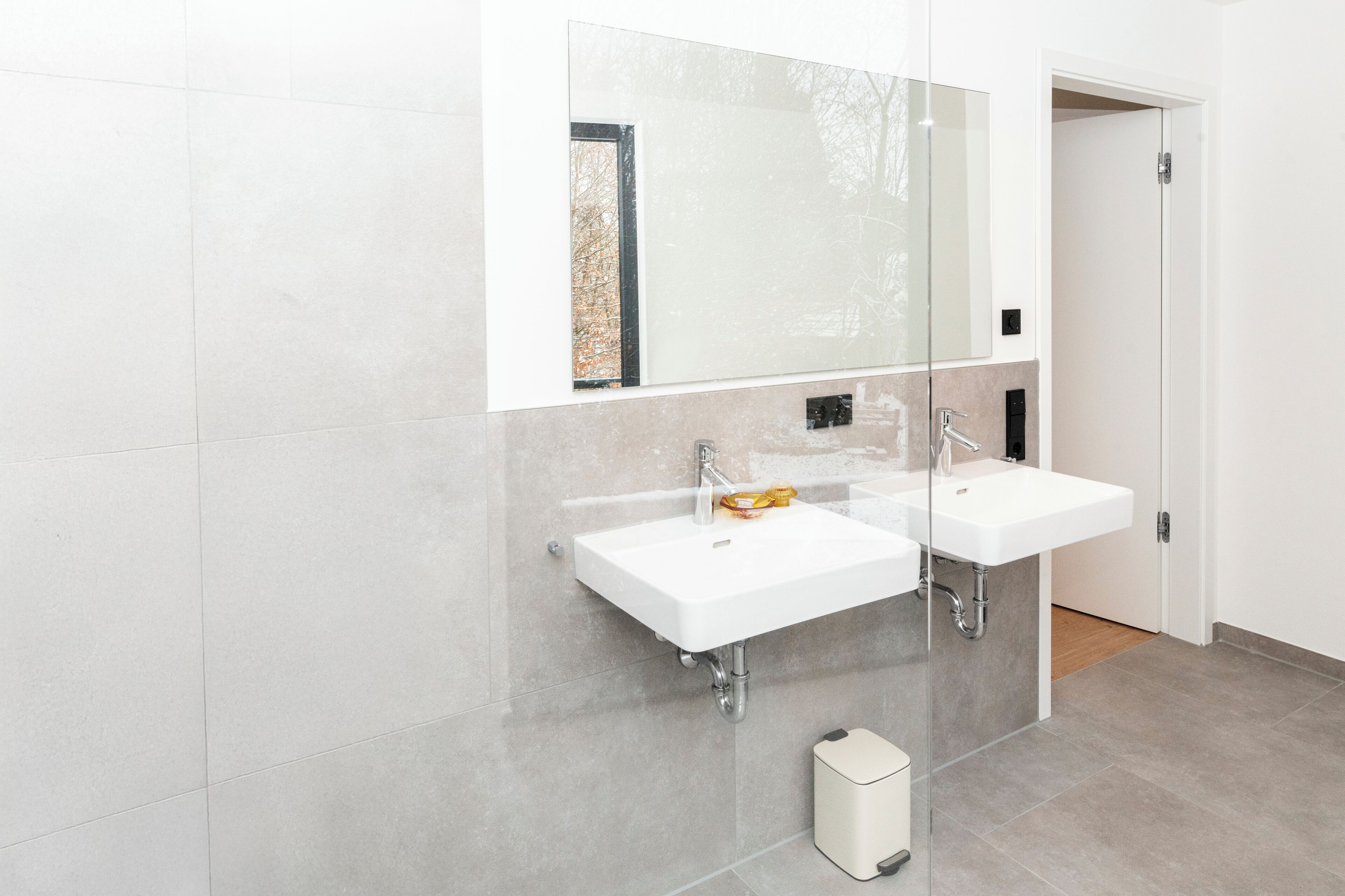 A modern bathroom with two white sinks, a large mirror, and a small trash bin.