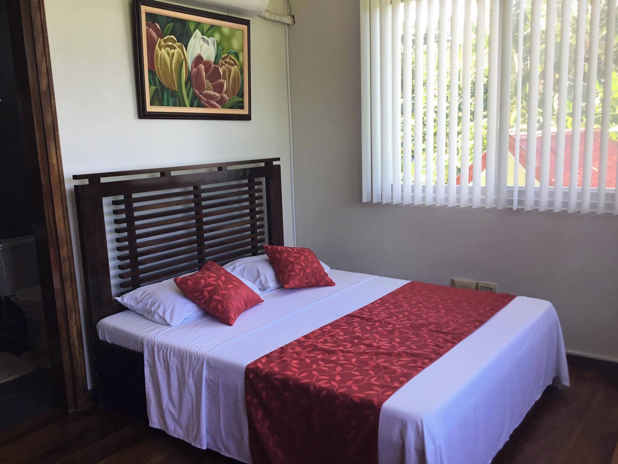 A neatly made bed with white and red bedding, a wooden headboard, and a painting of flowers on the wall.