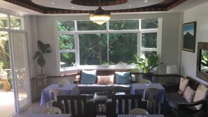 A dining area with tables covered in checkered tablecloths, a sofa with cushions, and a large window with a view of greenery.