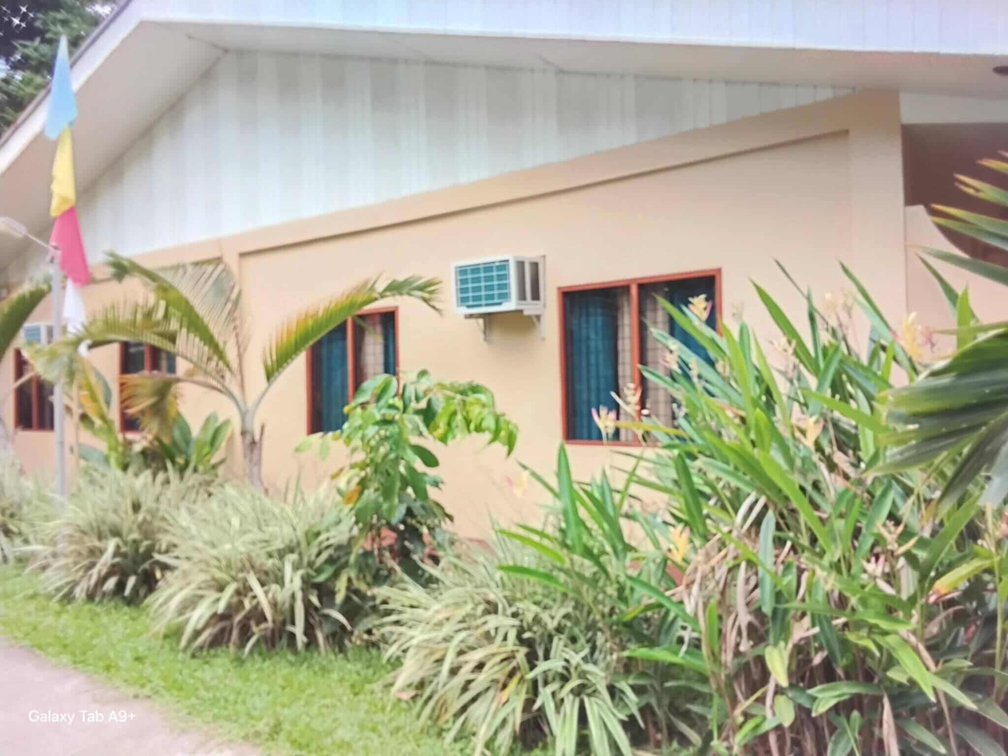 A building with a beige exterior, two windows with curtains, an air conditioning unit, and lush greenery in front.