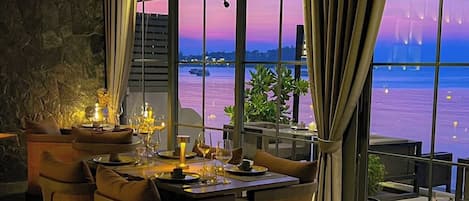 A restaurant with a view of the water, tables set with wine glasses and plates, and a stone wall.