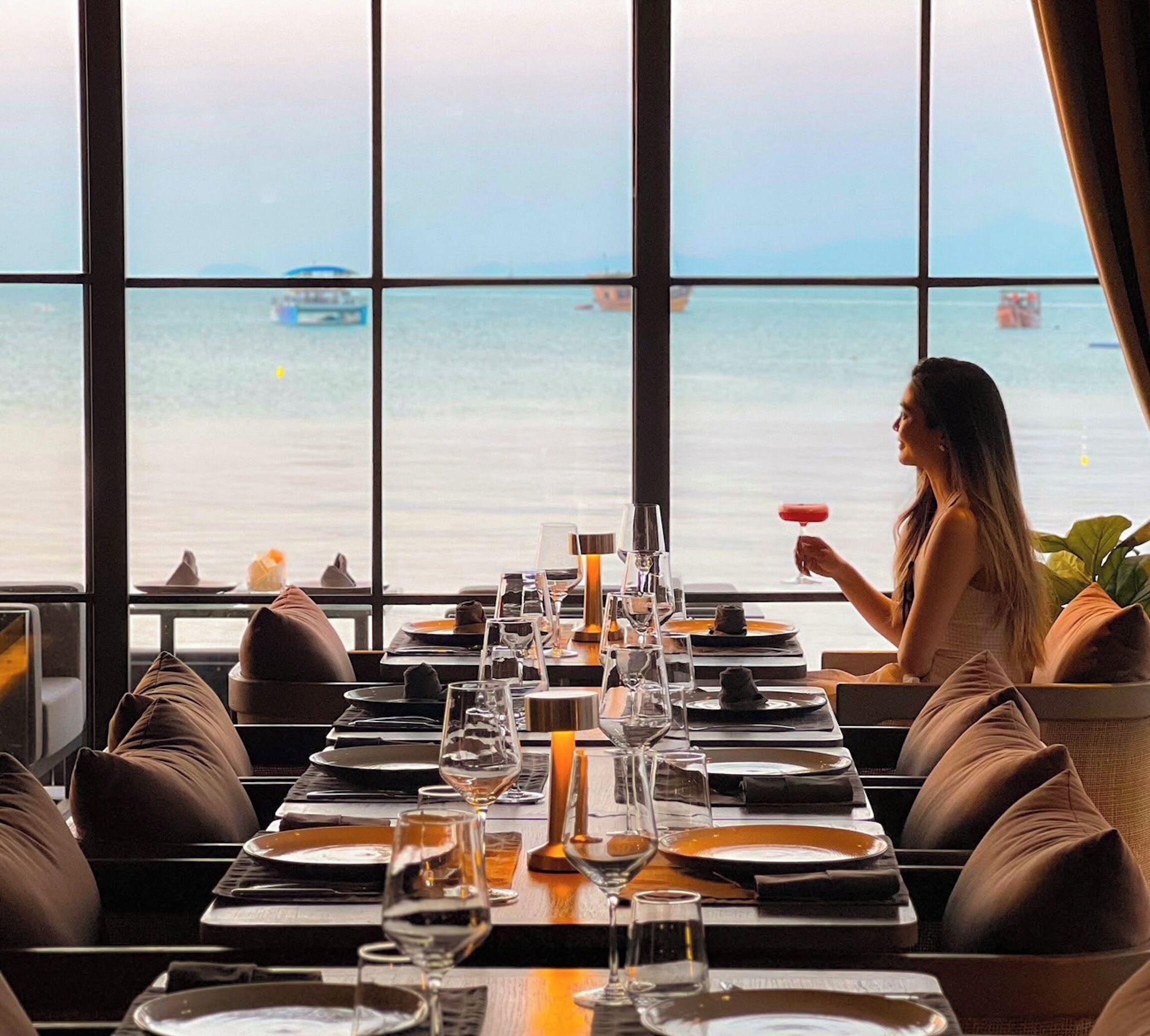 Een eetruimte met gedekte tafels, grote ramen met uitzicht op zee, en iemand die een drankje vasthoudt.