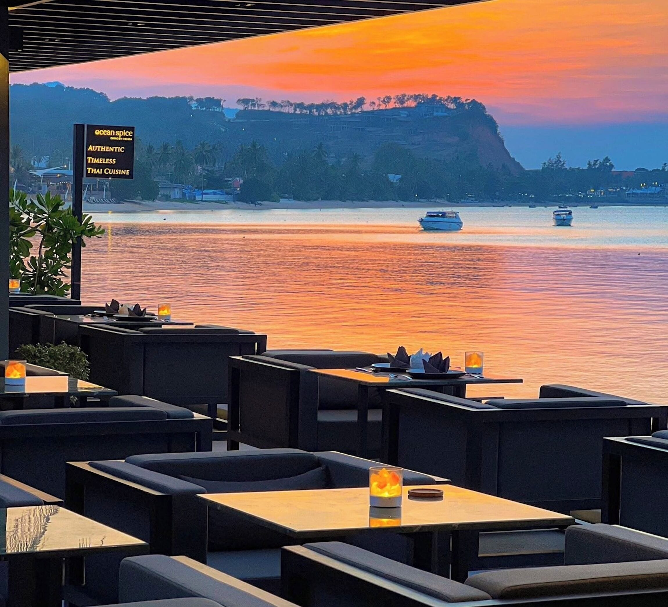 A restaurant with outdoor seating overlooking a body of water, a signboard, and a view of the water and distant hills.