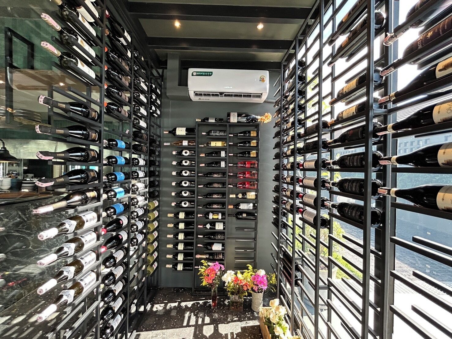 Une cave à vin avec des rangées de bouteilles de vin alignées sur des étagères, un appareil de climatisation et un petit arrangement floral posé sur le sol.