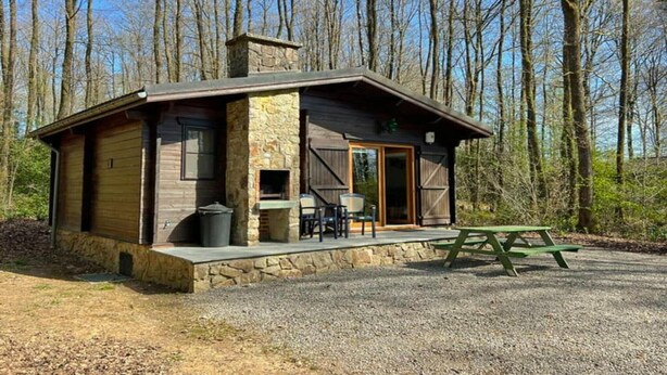 A cabin with a stone exterior, a chimney, a patio with chairs and a table, and a forested backdrop.