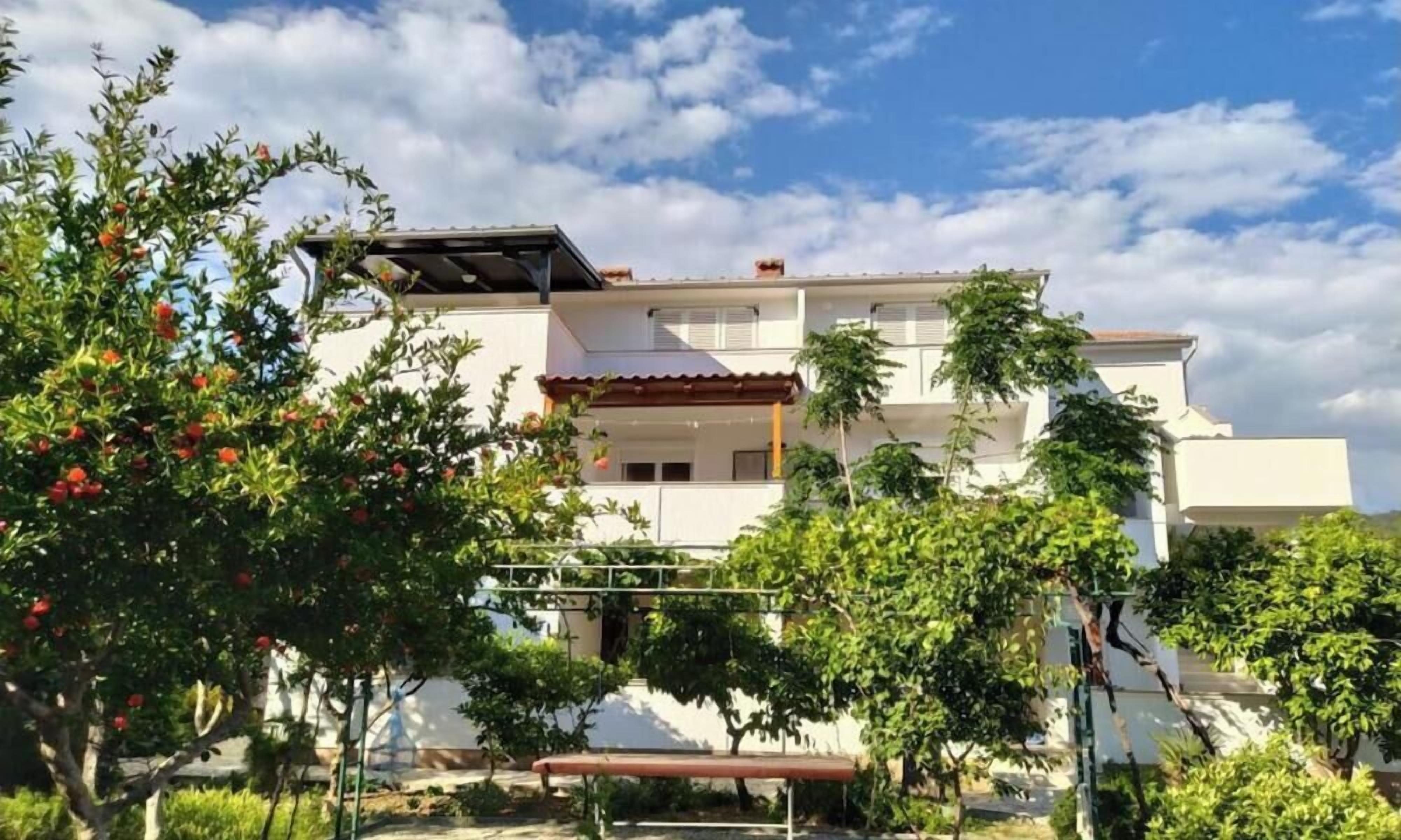 A two-story building with a flat roof, a balcony on the upper floor, and a garden area with trees and shrubs.