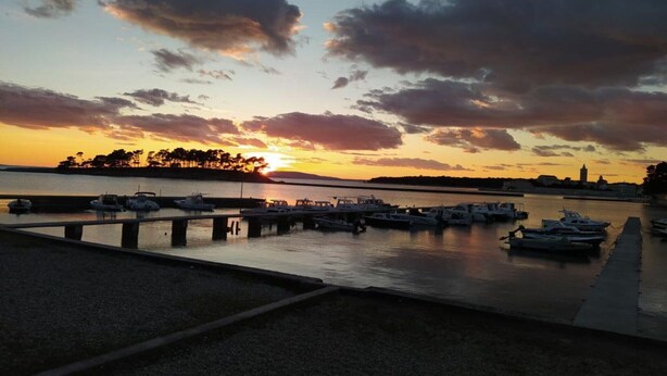 Una marina al atardecer con barcos amarrados, una superficie de agua tranquila y una costa lejana con árboles y un edificio.