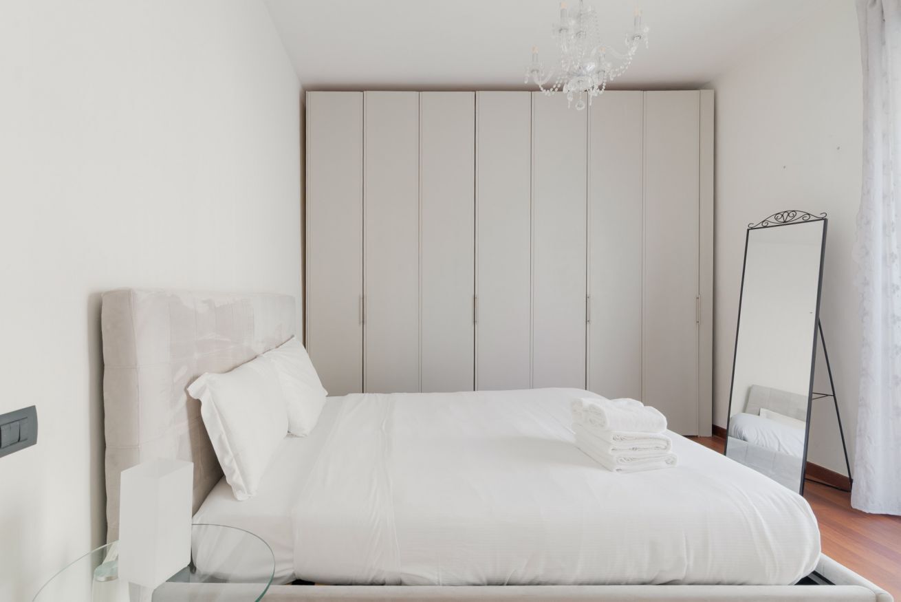 Een slaapkamer met een bed, nachtkastje, spiegel, kledingkast en een kroonluchter.