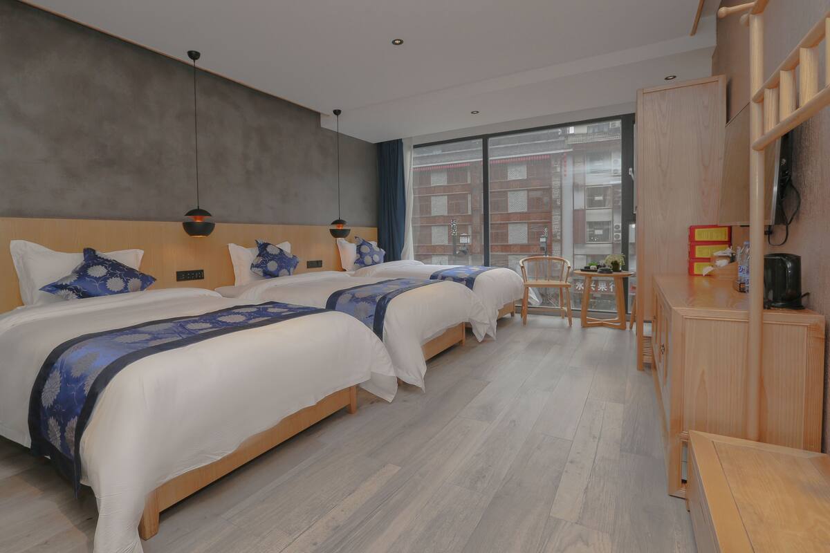 A modern hotel room with two beds, a wooden headboard, a small table, and a view of buildings through large windows.