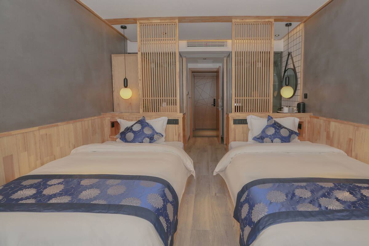 A hotel room with two beds, wooden headboards, a small kitchenette, and a bathroom visible through a door.