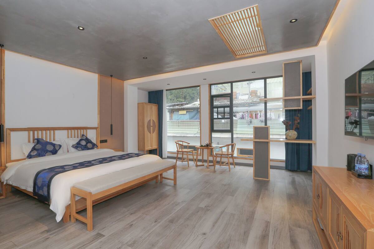 A modern hotel room with a large bed, wooden furniture, a dining area, and large windows offering a view of the outside.