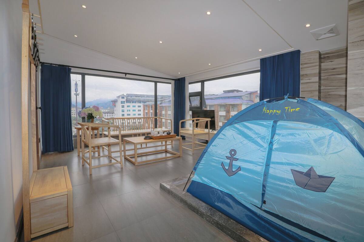 A spacious room with a large window, a blue tent, wooden furniture, and a view of buildings outside.