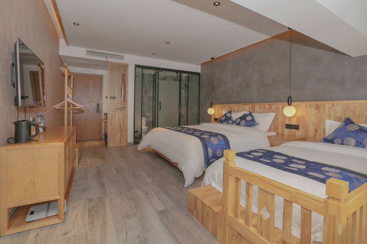 A modern hotel room with two beds, a wooden dresser, a mirror, and a bathroom visible through a glass door.