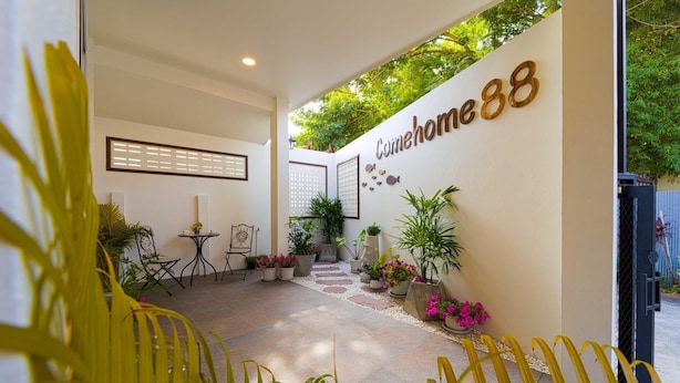 Ein moderner Hotel Eingangsbereich mit einem Schild, auf dem "Comerowhome88" steht. Draußen befinden sich Sitzbereiche mit Tischen und Stühlen, umgeben von Topfpflanzen, und ein Weg führt zum Eingang.