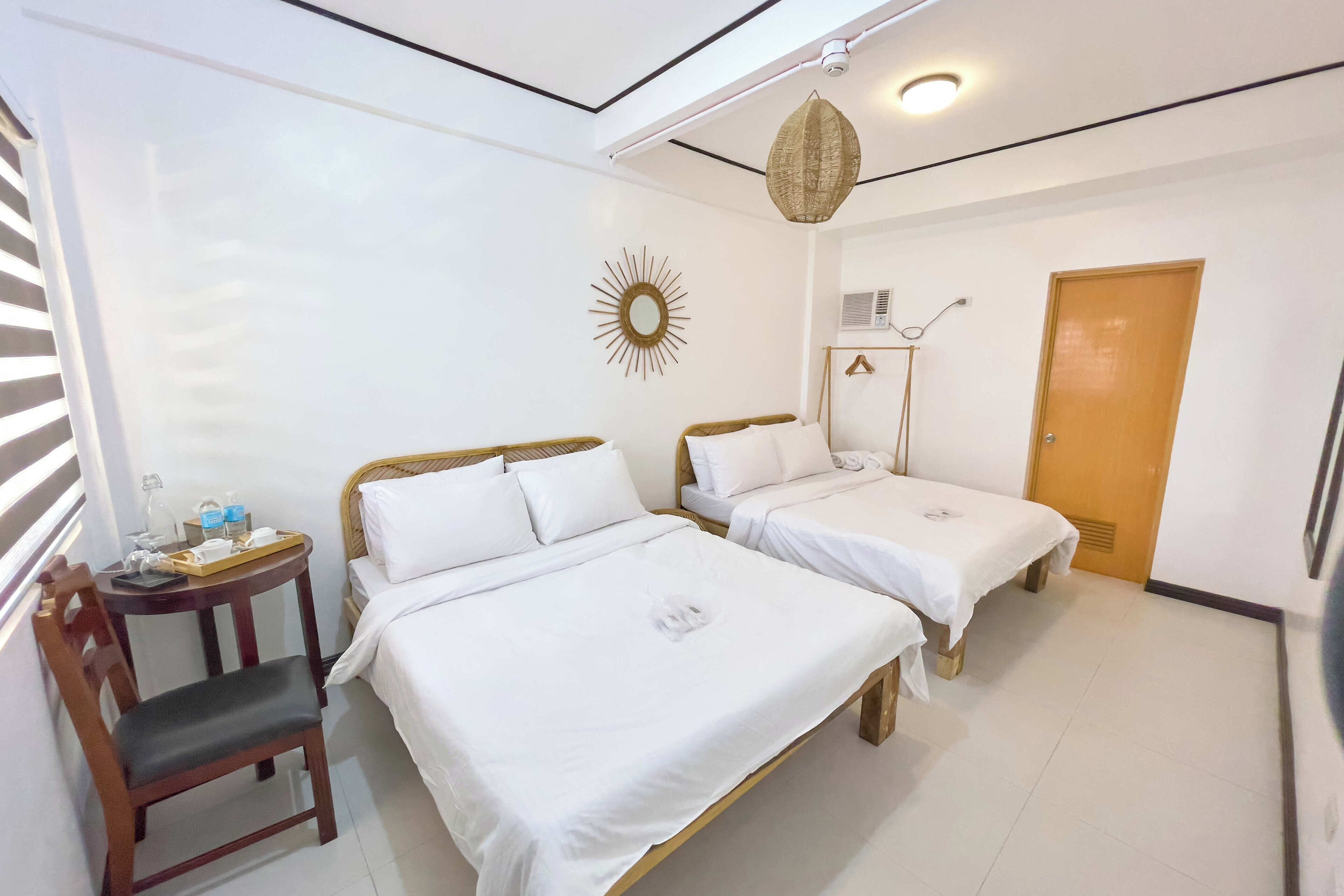 A hotel room with two beds, a small table with toiletries, a chair, a sunburst mirror, and a wooden door.
