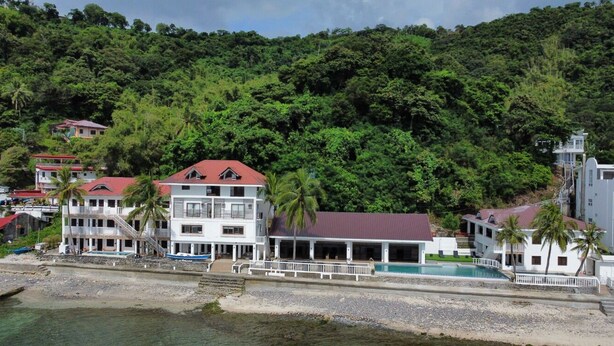 A beachfront property with multiple buildings, including a large white building with a red roof and a smaller structure with a white roof. There is a pool area with a white fence and palm trees. - Salacia Beachfront Resort (Bauan)