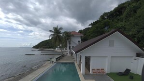 A poolside view of a resort with a clear view of the ocean, a small building with a red roof, and a grassy area with lounge chairs. - Salacia Beachfront Resort (Bauan)