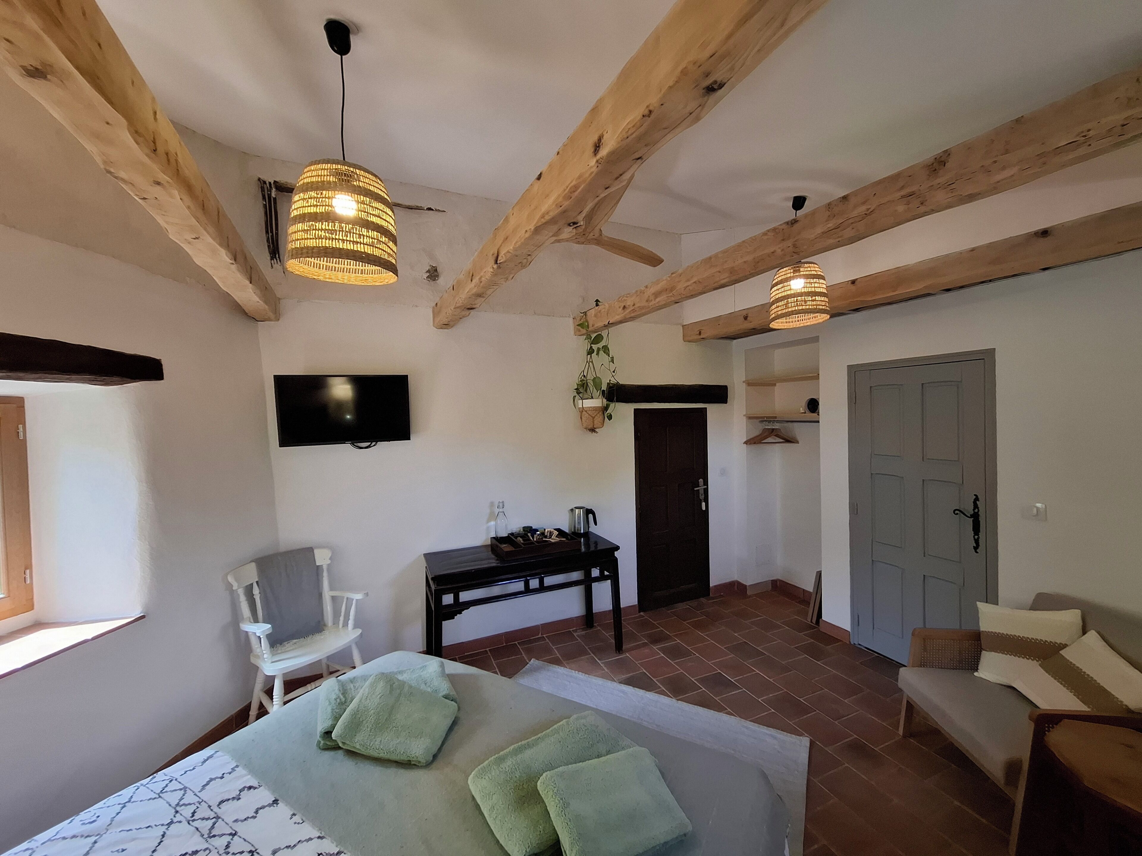 Een kamer met zichtbare houten balken, een bed met groene handdoeken, een televisie aan de muur, een tafel met een fles en waterkoker, en een deur met een hangende plant.